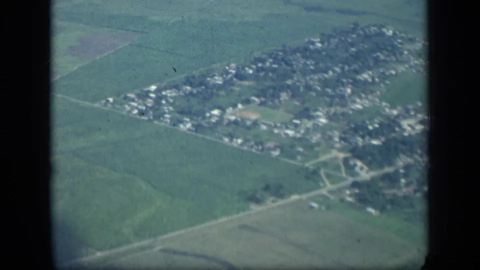 Aerial view over a small New York town surrounded by farmland, with roads, houses, and open fields visible in soft 8mm color.
