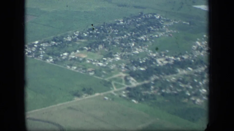 1957 New York home-movie aerial view of a small town among green fields, with roads and houses seen through an airplane window.
