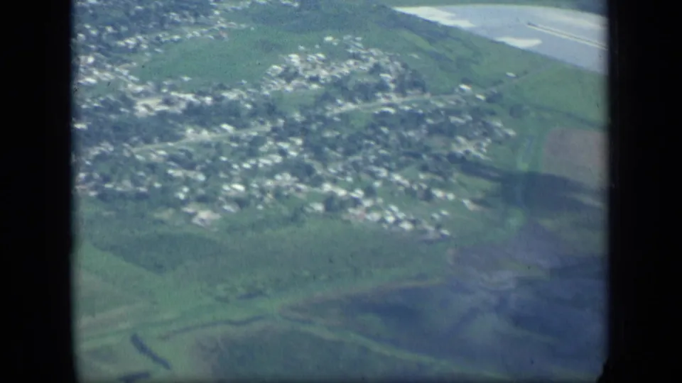 1957 New York airplane-window view of a town and fields, with a pale water or shoreline area visible near the upper edge.