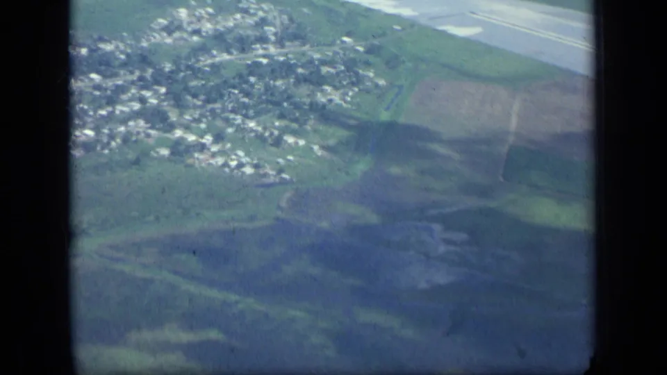 1957 New York aerial home-movie frame showing a small town, surrounding farmland, darker lowland vegetation, and distant pale water.