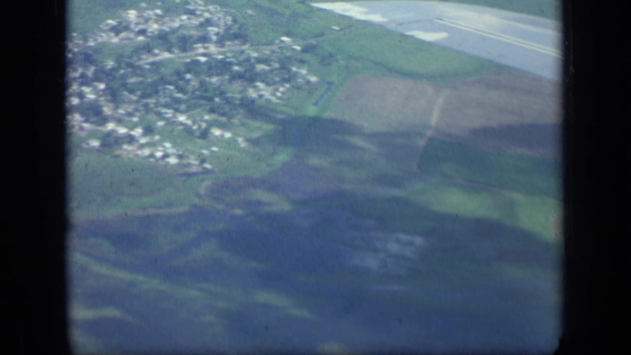 Aerial view of fields, hills, small buildings, and part of a runway seen through dark film-frame borders.