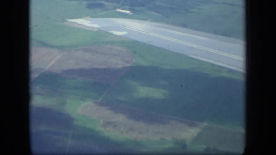 1957 New York aerial frame with a runway, fields, wooded terrain, and soft shadow patterns seen from a moving airplane.