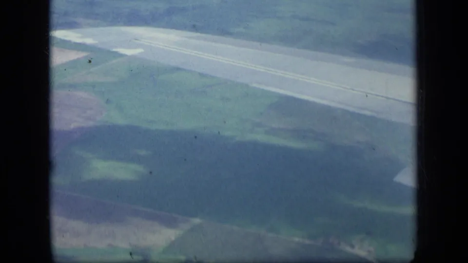 1957 New York 8mm airplane-window view showing a runway across rural fields and wooded hills with visible film-frame borders.