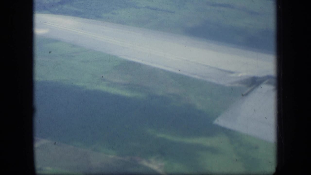 Airplane wing above pale green land and water or shoreline, framed by dark film gate edges.