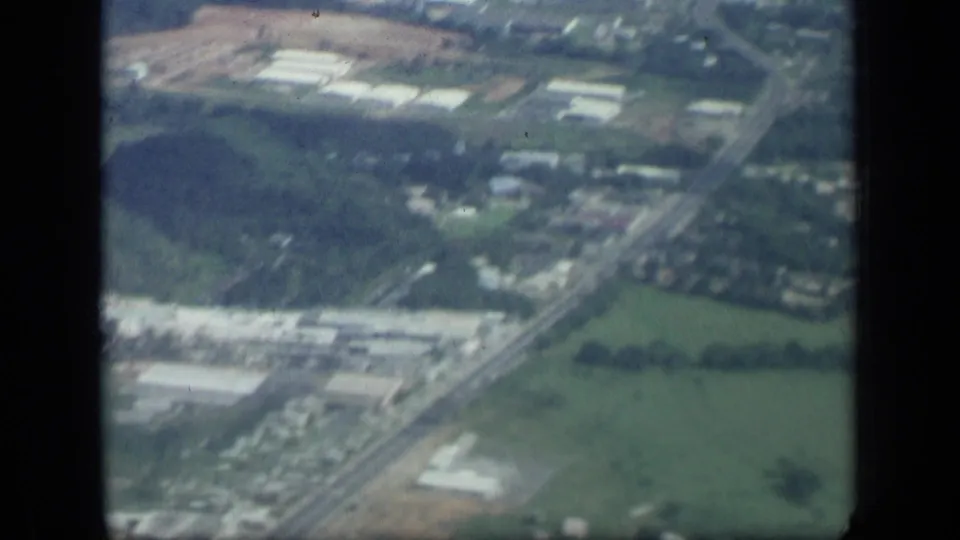 1957 New York aerial home-movie frame with a road running past green fields, clustered buildings, and wooded terrain.