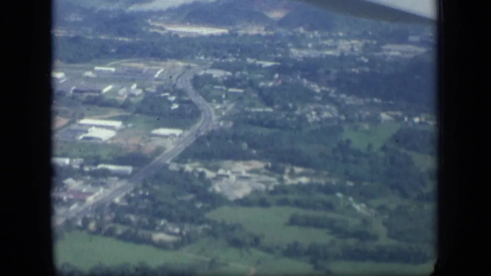 1957 New York home-movie frame showing a small-town landscape of fields, roads, and buildings viewed from an airplane window.