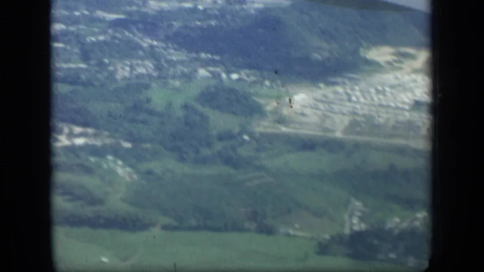 Soft 8mm aerial frame from 1957 New York showing green land, roads, scattered buildings, and a pale developed area below.