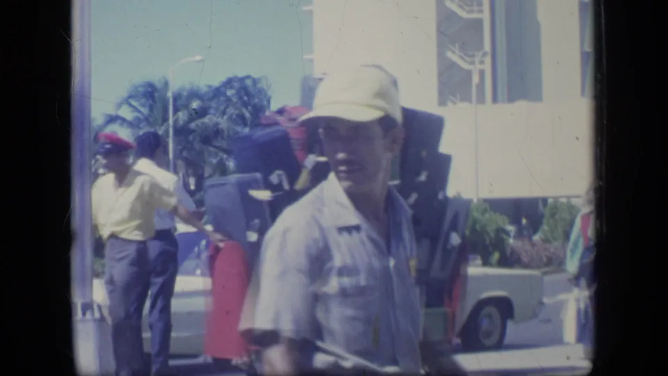 1957 New York travel scene with a man in a light cap near luggage, other people, and a vehicle in bright outdoor light.