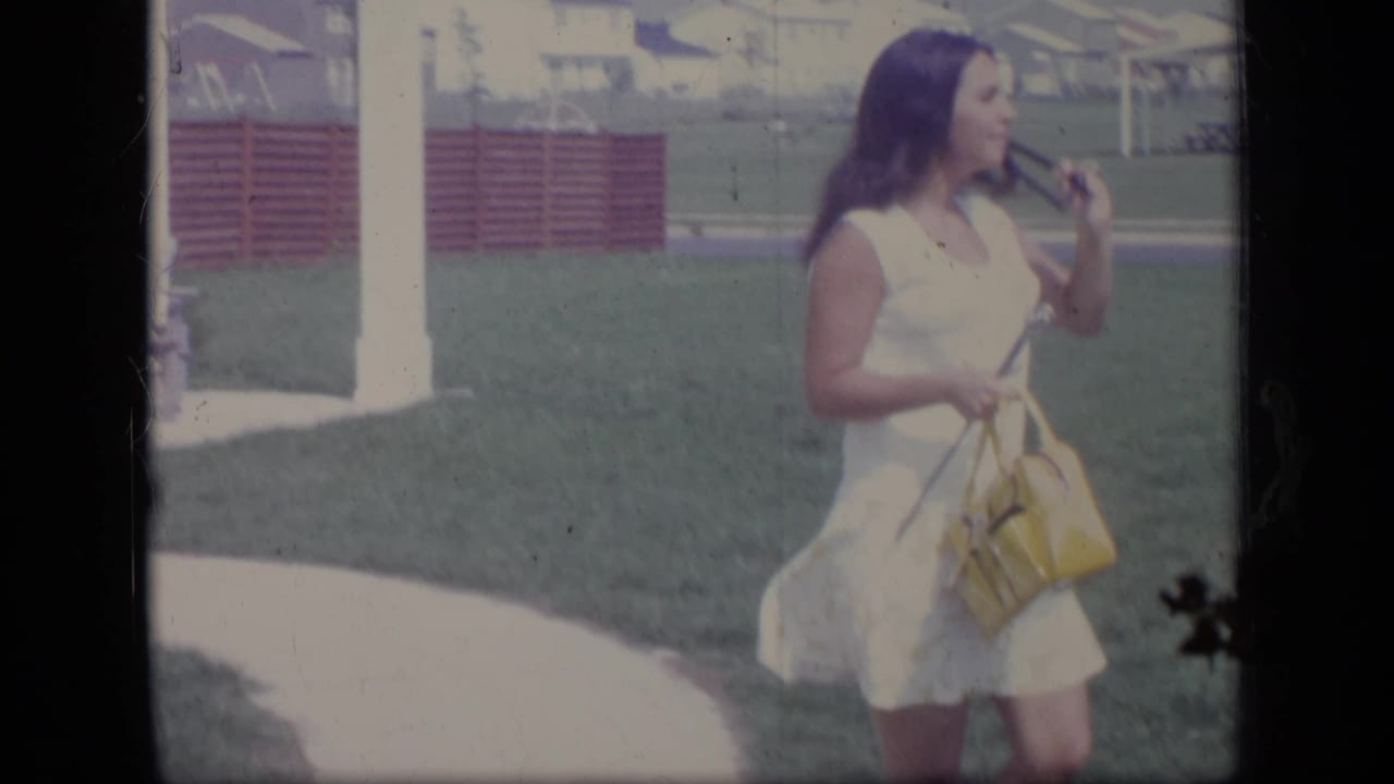 Woman in a white dress walks on a lawn carrying a yellow handbag and long tool.