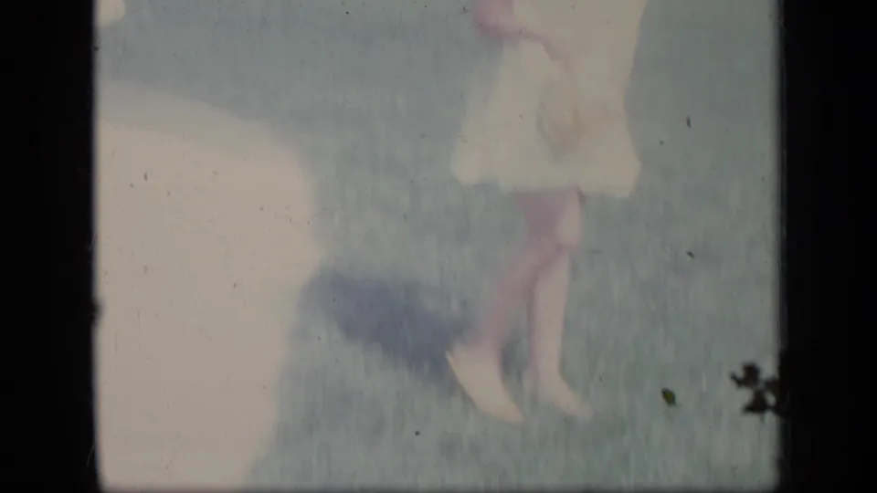 A washed-out 8mm frame shows the woman’s white dress, legs, and shadow crossing the grass in the suburban yard.
