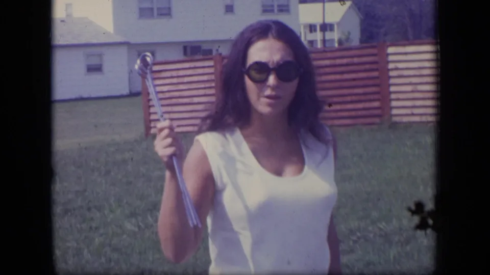 In the New Jersey yard, the woman faces the camera wearing dark sunglasses and holding a long metal tool upright beside her shoulder.