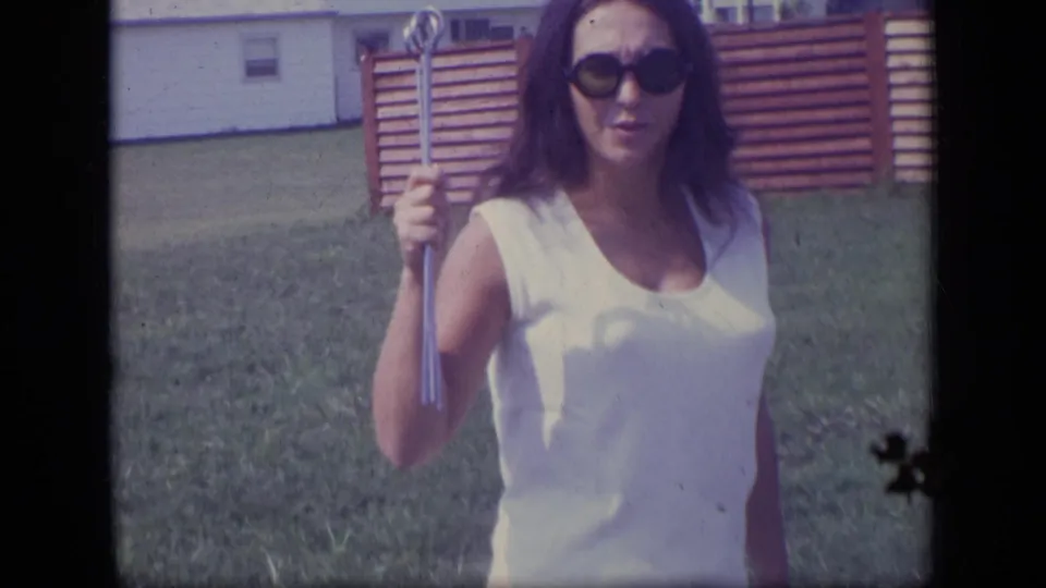 The woman stands on the lawn in a white dress and dark sunglasses, holding the long tool near her shoulder with fence and houses behind her.