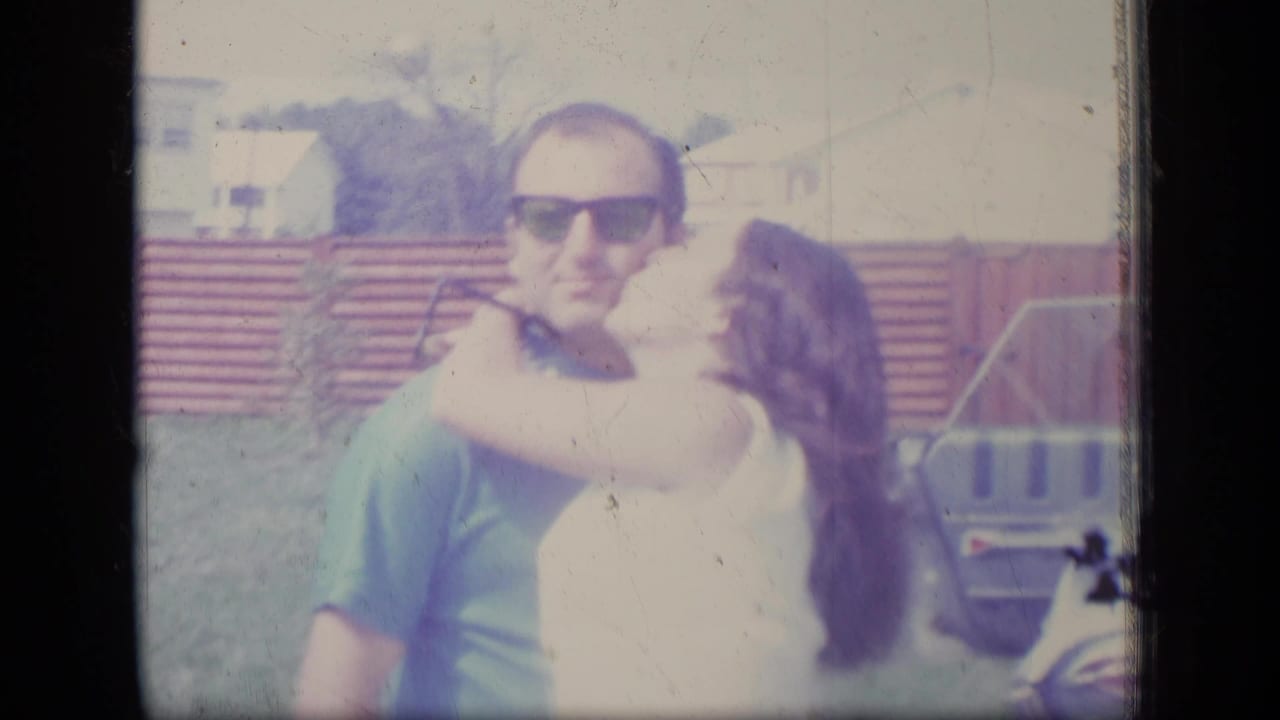 Man in sunglasses embraces a woman in a backyard near a fence.