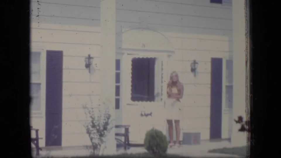 A woman stands outside the front door of a white suburban house with dark shutters in 1970 New Jersey home-movie footage.