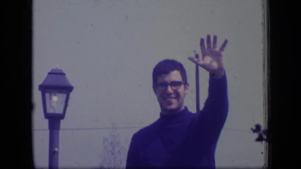 A man wearing glasses and a dark turtleneck waves toward the camera near an outdoor lamp in 1970 New Jersey footage.