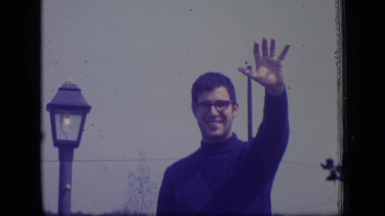 In this 1970 New Jersey 8mm frame, an adult man in glasses raises his hand in a cheerful wave near a street lamp and pale wall.