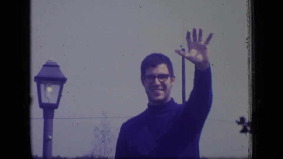 In this 1970 New Jersey 8mm frame, an adult man in glasses raises his hand in a cheerful wave near a street lamp and pale wall.