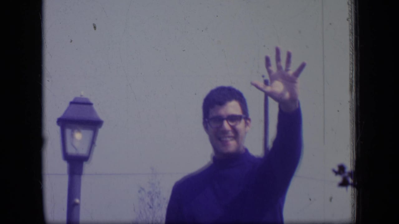The man continues waving in the 1970 New Jersey home-movie frame, with a street lamp and plain wall behind him.