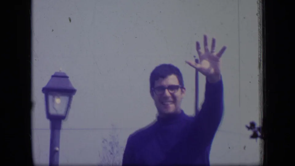 The man continues waving in the 1970 New Jersey home-movie frame, with a street lamp and plain wall behind him.