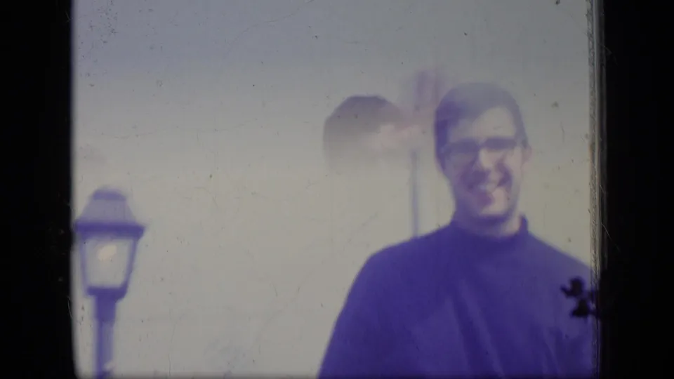 A soft and overexposed 8mm frame shows the smiling man in glasses near the lamp post as the camera or film shifts.