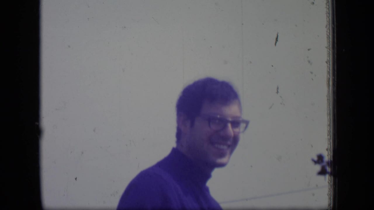 In supported 1970 New Jersey home-movie footage, a man in glasses and a blue turtleneck smiles toward the camera against a pale outdoor background.