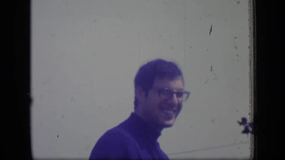 In supported 1970 New Jersey home-movie footage, a man in glasses and a blue turtleneck smiles toward the camera against a pale outdoor background.
