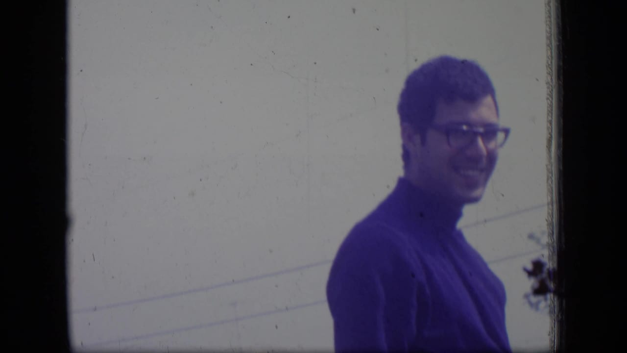 Man in glasses and a blue turtleneck smiles in profile outdoors.