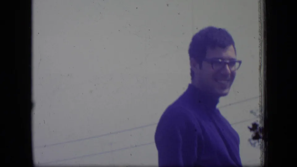 A man in glasses and a blue turtleneck smiles in profile in the 1970 New Jersey home-movie frame, with faint outdoor lines behind him.