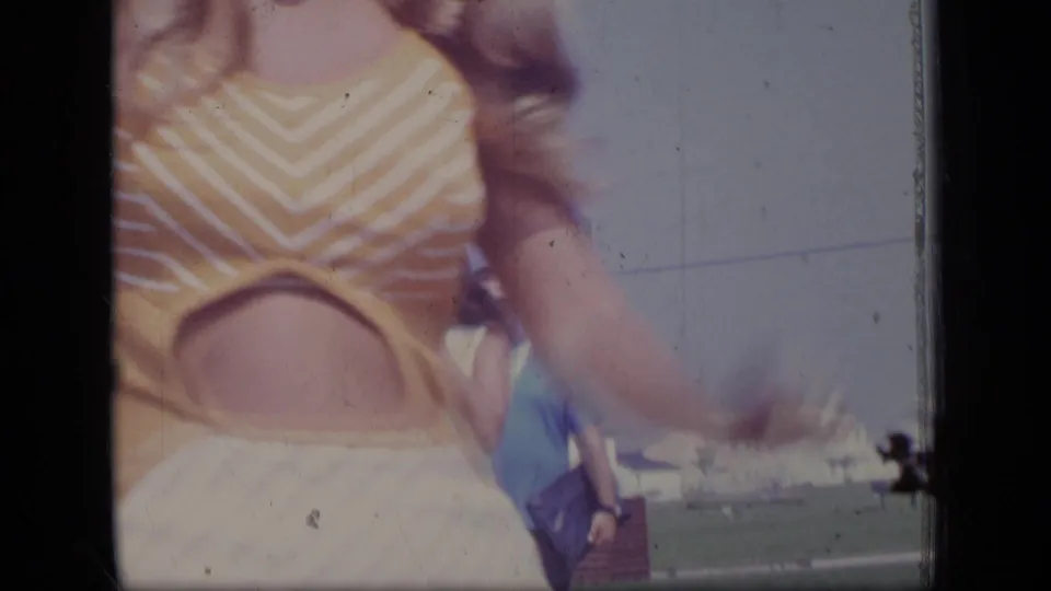 A woman in a yellow and white striped top moves close past the lens, with lawn and distant outdoor details visible in the 1970 New Jersey footage.