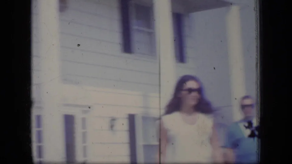A woman in a white dress and sunglasses walks outdoors near a light-colored house, with another adult partly visible in the 1970 New Jersey frame.