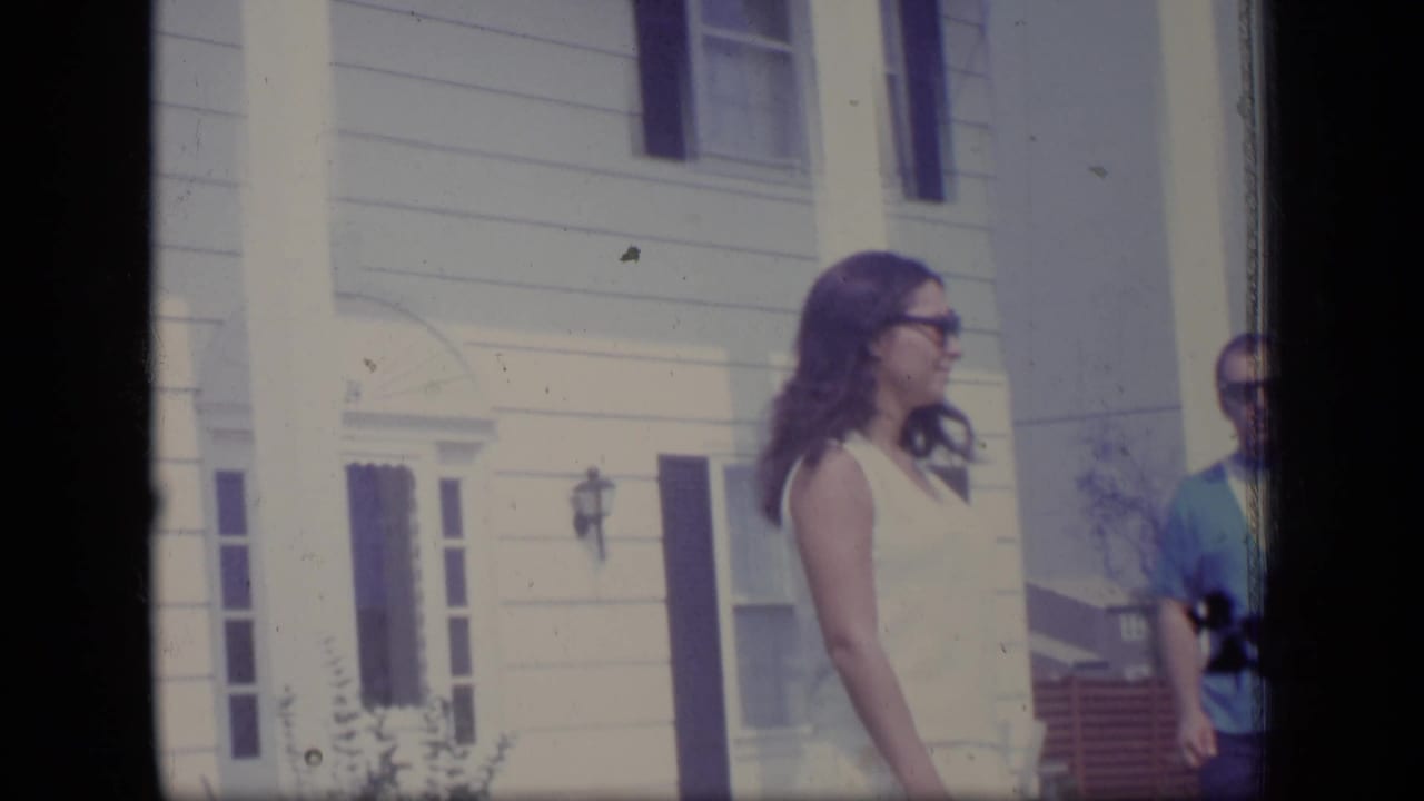 Woman in sunglasses stands outside a white clapboard house as a man is partly visible nearby.