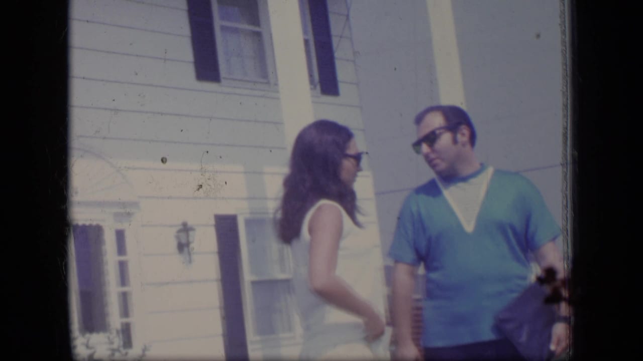 1970 New Jersey home-movie frame of a man in a blue V-neck shirt and a woman in white conversing outside a white clapboard house.