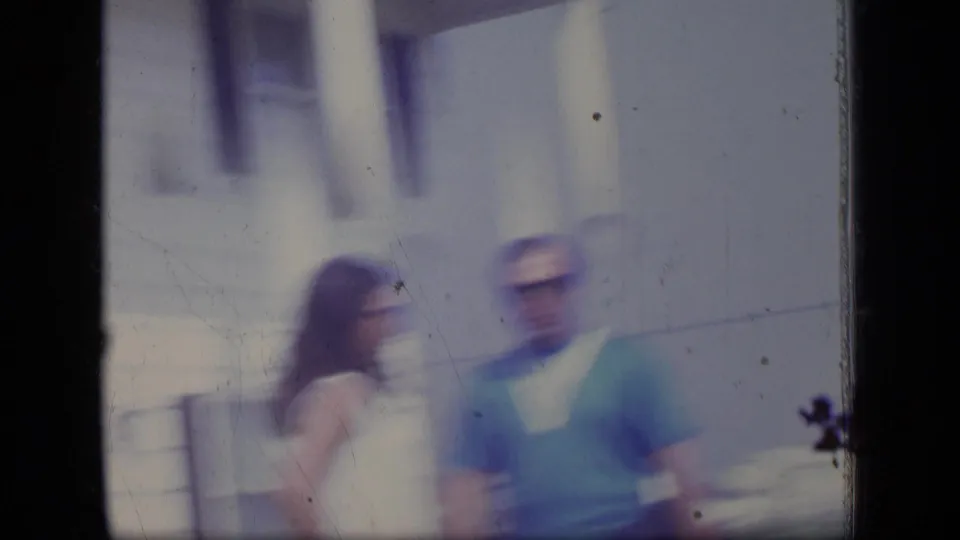 Soft, motion-blurred 1970 New Jersey home-movie frame showing the couple shifting position outside the clapboard house.