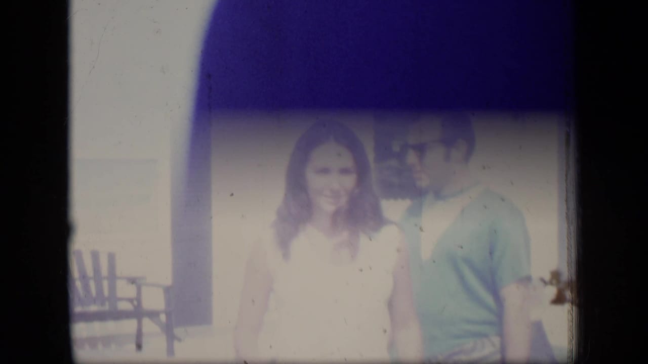 Woman in a white sleeveless dress and man in a blue t-shirt stand outside a white house.