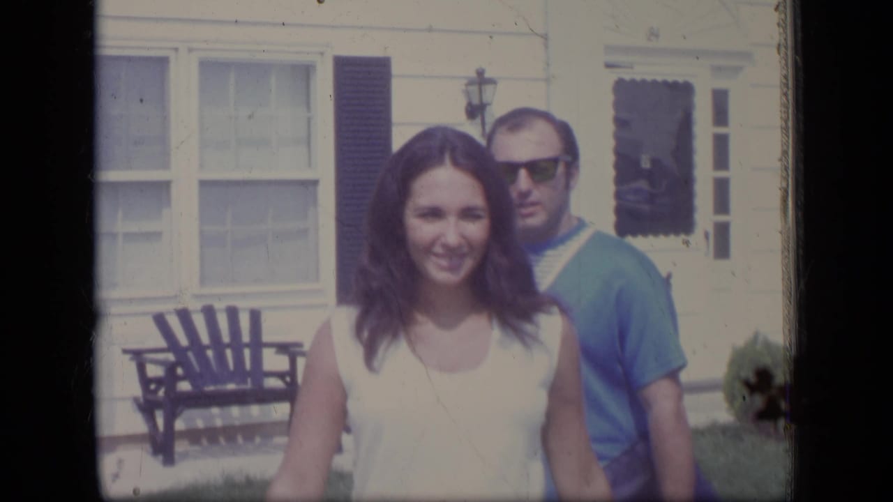 A smiling woman in a white sleeveless dress stands in front of a man wearing sunglasses outside a New Jersey suburban home in 1970.