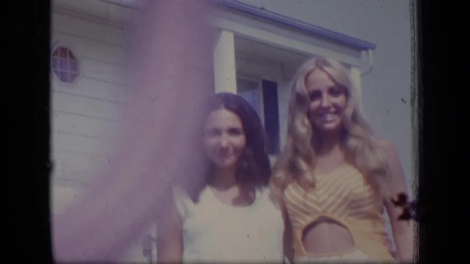 Two women stand beside each other outside a New Jersey house in 1970, one wearing a white sleeveless dress and the other a striped yellow cutout dress.