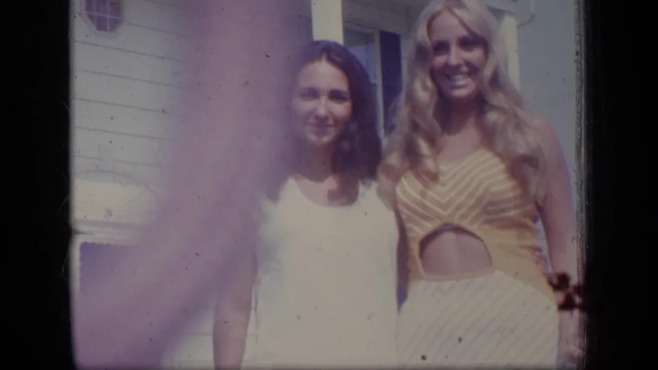 Two women pose outside a white-sided New Jersey home in 1970, with visible 8mm color shift and flare across the frame.