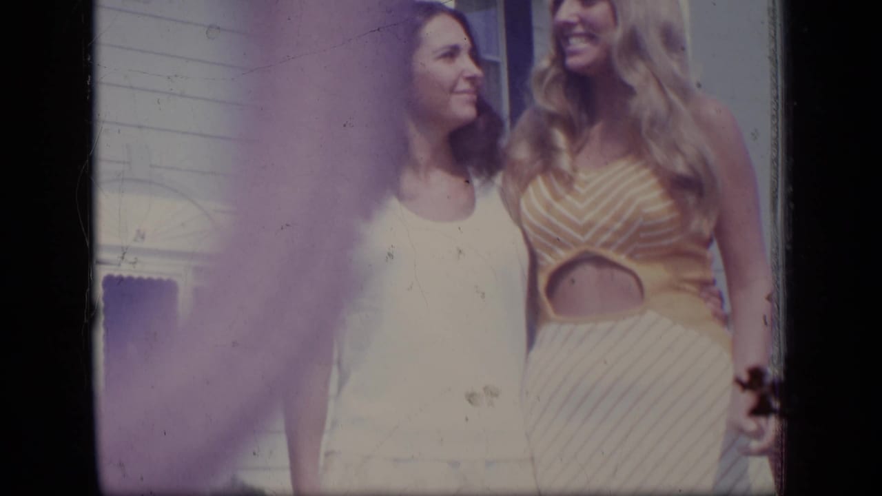 Two women smiling together outdoors beside a white house.