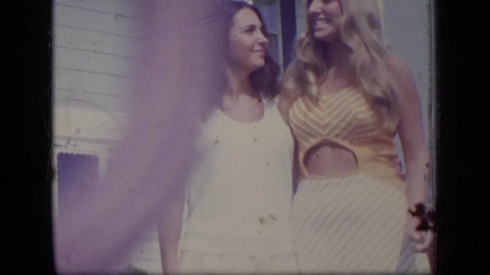 Two women in 1970s New Jersey home-movie footage stand close together in a sunny yard beside a white house.