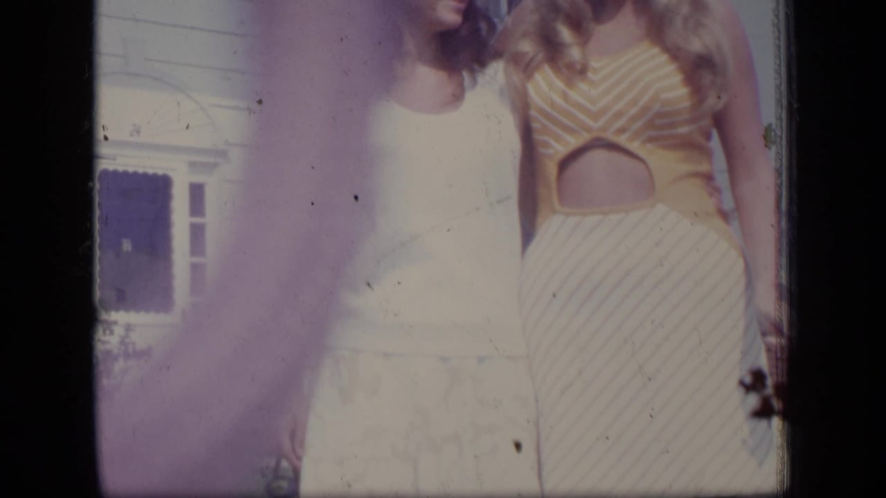 The camera frames two women from the shoulders to knees, with a yellow striped outfit and white dress visible in bright outdoor light.