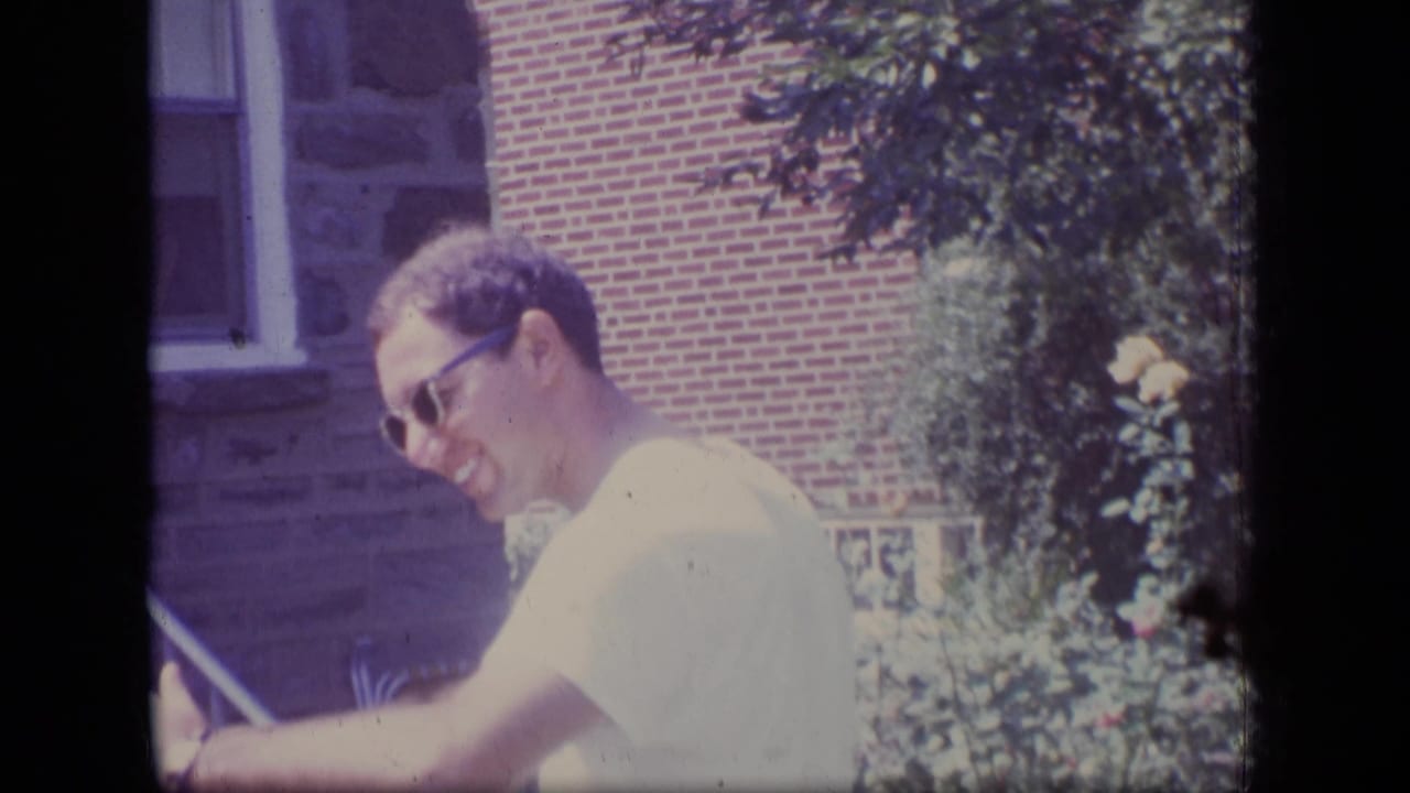 Man in sunglasses laughs outdoors near a brick and stone house.