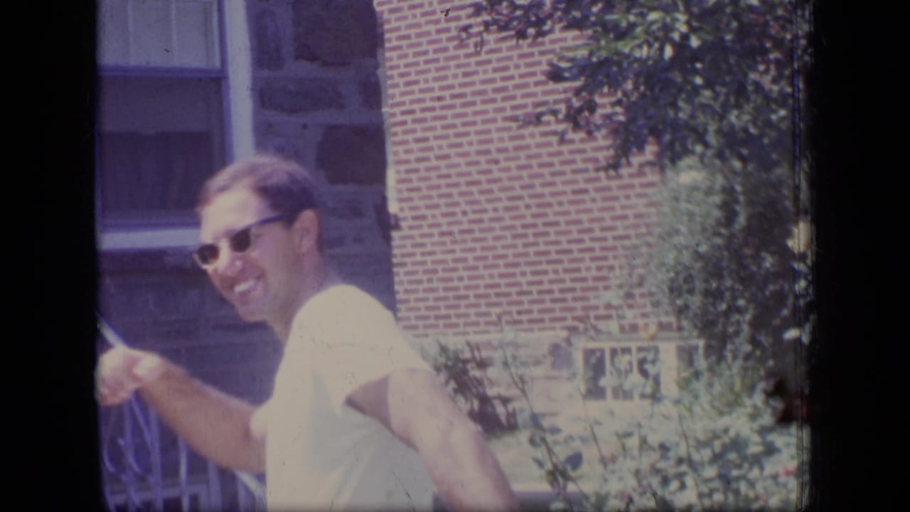 The man smiles broadly while turning near the brick and stone house exterior, with shrubs and windows in the background.