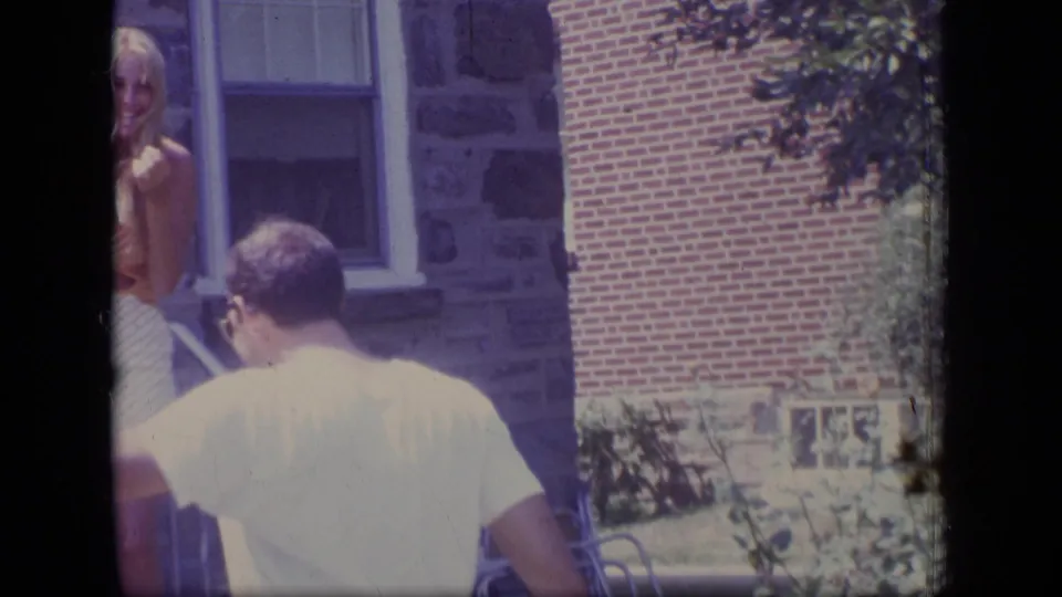 The man is seen from behind near a railing while a partly visible woman stands at the edge of the frame outside the house.