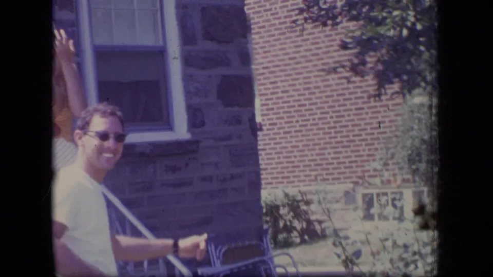 The smiling man in sunglasses stands near a porch railing with the brick-and-stone house exterior behind him.