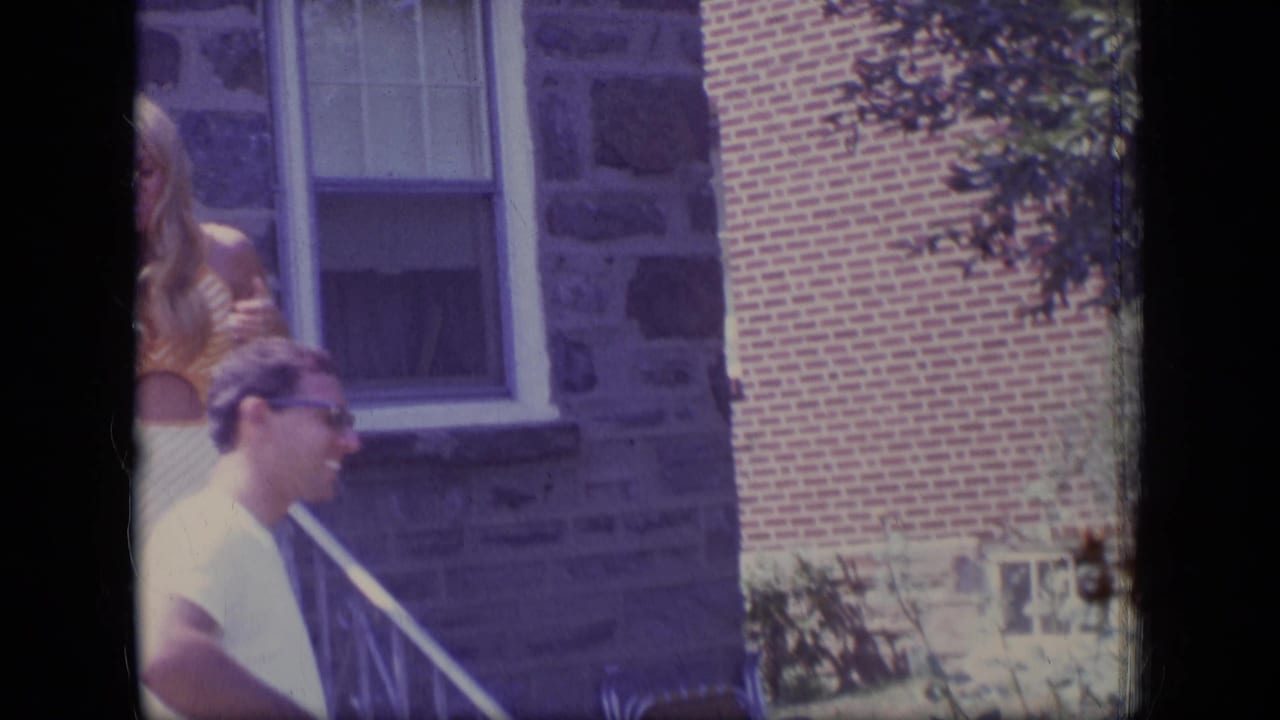 1970 New Jersey 8mm frame showing a man in a white shirt and sunglasses near a blonde woman on porch steps outside a stone and brick house.