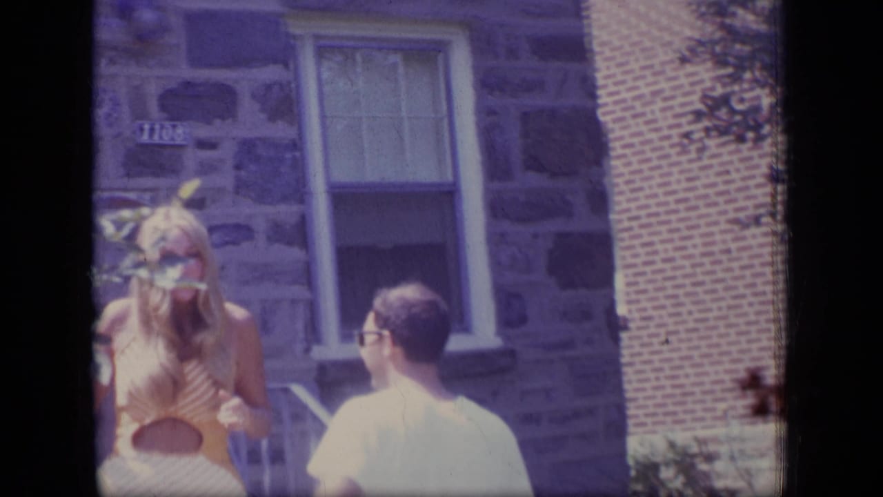 1970 New Jersey home-movie frame with a blonde woman in a sleeveless dress facing a man in sunglasses beside porch steps and stonework.