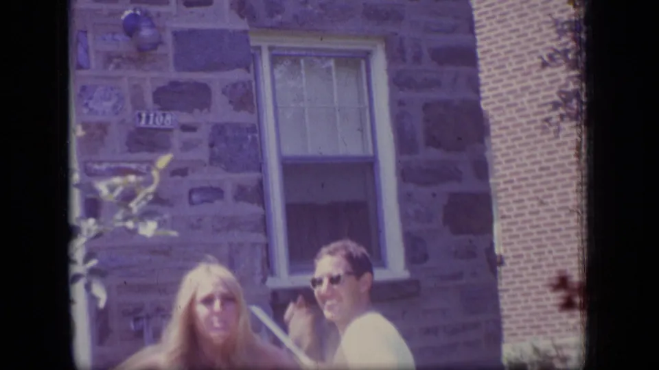 1970 New Jersey 8mm frame showing the woman and man turning toward the camera outside a stone-and-brick residential exterior.