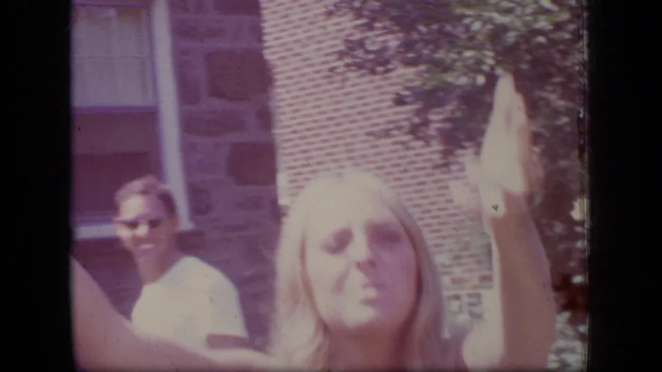 1970 New Jersey home-movie frame with a blonde woman close to the camera raising an arm as a man in sunglasses stands near the house behind her.