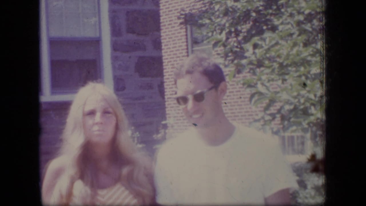 Woman in striped top and man in sunglasses stand outside a stone and brick building.