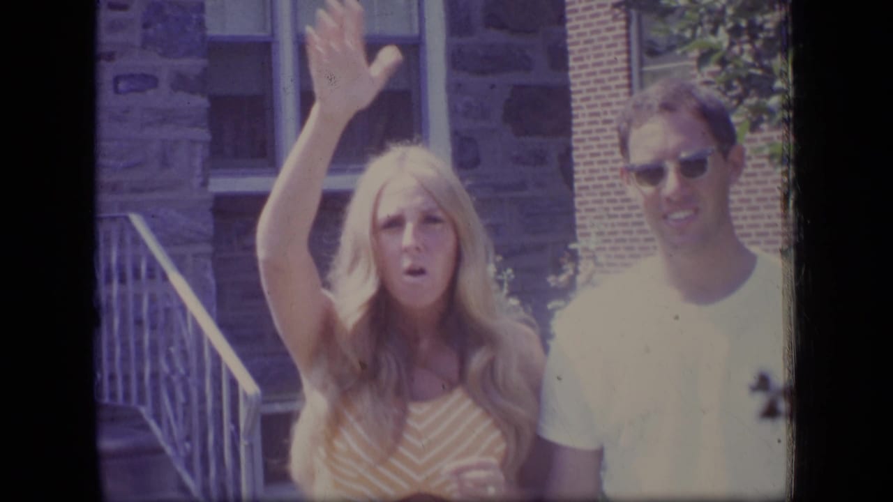The woman lifts one arm in a wave while standing beside a man outside a stone building in the 1970 New Jersey clip.